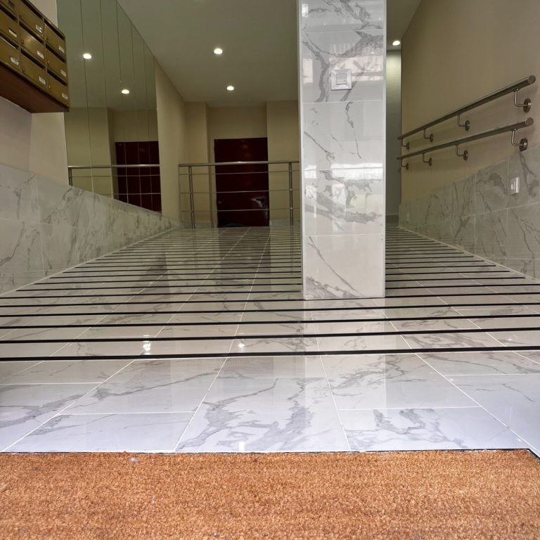 Rehabilitación comunitaria Vestíbulo comunidad de vecinos con suelo de mármol pulido con líneas oscuras y pared brillante, interior moderno y minimalista.